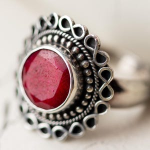 May include: A silver ring with a large, round, red gemstone at the center. The ring has an ornate design with a beaded border and a series of infinity-shaped accents around the gemstone.