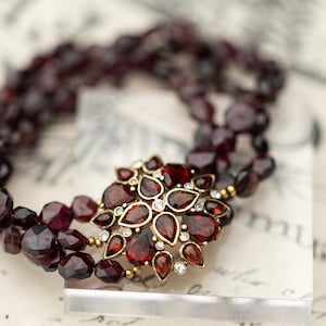 May include: A multi-strand bracelet composed of deep red, faceted garnet beads. The centerpiece is a floral-shaped gold-tone metal clasp featuring teardrop-shaped garnet stones and small, clear, round accent crystals arranged in a cluster.