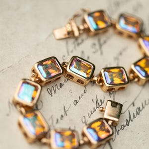May include: A gold-toned link bracelet featuring a series of rectangular, faceted amber-colored stones with iridescent highlights. The bracelet is constructed with individual metal settings for each stone and includes a secure box-style clasp closure.