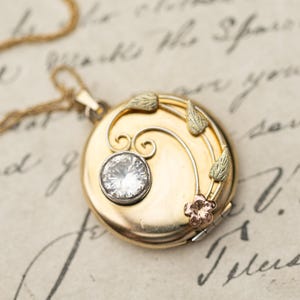 Könnte beinhalten: Ein runder Goldmedaillon-Anhänger an einer dünnen Goldkette mit einem einzelnen klaren, runden Edelstein in einer silberfarbenen Fassung, verziert mit gravierten Blattmotiven und einer kleinen fünfblättrigen Blume in kontrastierendem Roségold.