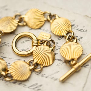 May include: A gold-toned bracelet featuring a series of interconnected shell-shaped charms with ribbed detailing. The piece includes a circular toggle ring and a straight bar clasp, along with a small square tag attached near the toggle closure.