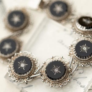 May include: A silver link bracelet featuring six circular black stone settings. Each stone is inlaid with a silver starburst design. The stones are framed by intricate silver filigree metalwork with a scalloped edge and a beaded border.