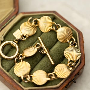 May include: A gold-toned bracelet featuring a series of interconnected shell-shaped links with a ribbed texture. The bracelet is secured with a toggle clasp consisting of a circular ring and a straight bar, and includes a small rectangular metal tag.