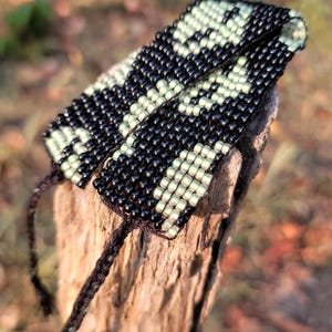 May include: A black and green beaded bracelet with a geometric pattern. The bracelet is tied with black string.