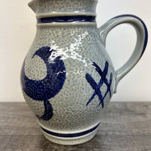May include: A gray ceramic pitcher with blue painted designs. The pitcher has a rounded body and a handle. The design includes a blue number "9" and a blue "X" pattern.