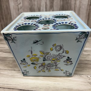 May include: A light blue ceramic flower frog with a square shape and a series of holes on the top. The sides are decorated with floral patterns in yellow, gray, and white. The edges are outlined in dark blue.