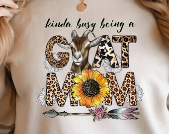 May include: A cream-colored sweatshirt with the text "kinda busy being a GOAT MOM" in black. The text is surrounded by a brown and black leopard print, a sunflower, and a white goat with brown spots.