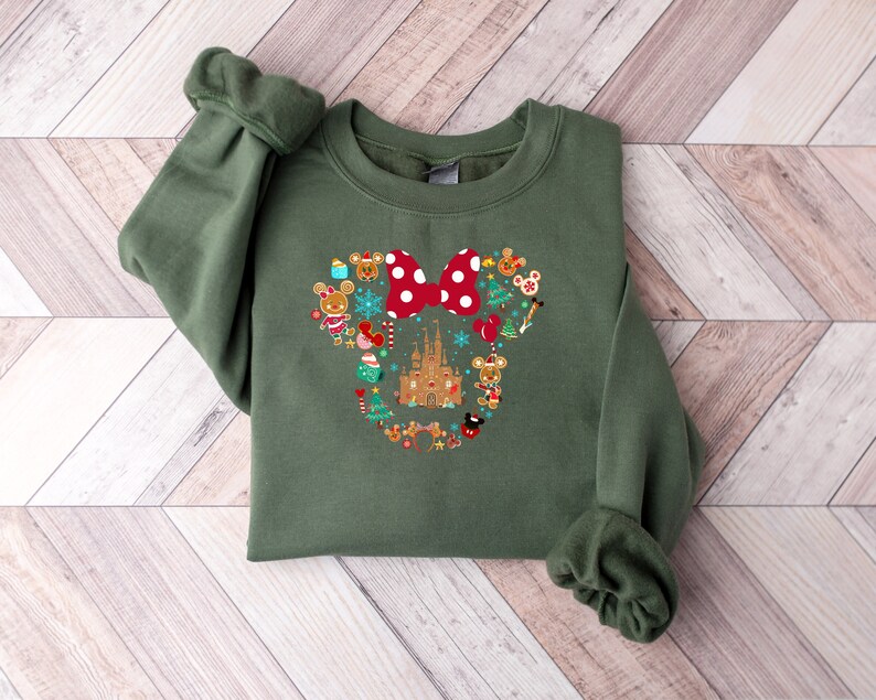 Pu&ograve; includere: Una felpa verde con un design a tema natalizio di Topolino. Il design presenta una testa di Topolino di pan di zenzero con un fiocco rosso, circondata da ornamenti di pan di zenzero, un castello di pan di zenzero e altri elementi a tema natalizio.