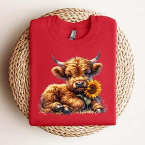 May include: Red t-shirt with a cartoon image of a brown Highland cow calf holding a yellow sunflower.