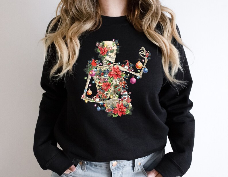 May include: Black sweatshirt with a skeleton wearing a Christmas tree costume. The skeleton is decorated with ornaments, lights, and poinsettias.