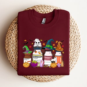 May include: A maroon t-shirt with a Halloween-themed design featuring three bottles with witch hats and the words "Diprivan", "Fentanyl", and "Ativan" on them.