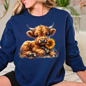 May include: A navy blue sweatshirt with a cute cartoon Highland cow holding a sunflower. The cow is brown and white with a fluffy mane.