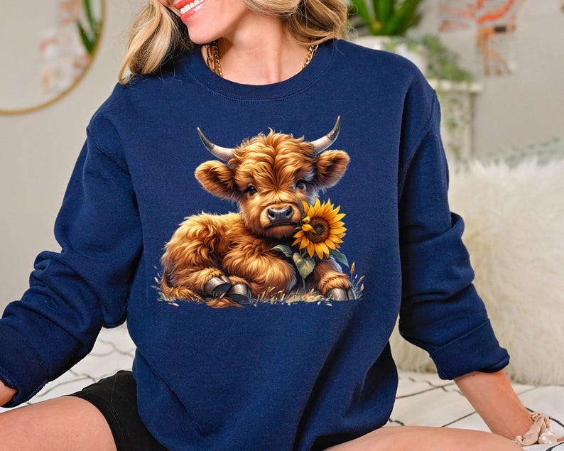 May include: A navy blue sweatshirt with a cute cartoon Highland cow holding a sunflower. The cow is brown and white with a fluffy mane.