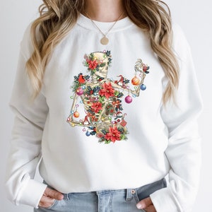 May include: White sweatshirt with a colorful illustration of a skeleton decorated as a Christmas tree. The skeleton is adorned with ornaments, lights, and poinsettias.