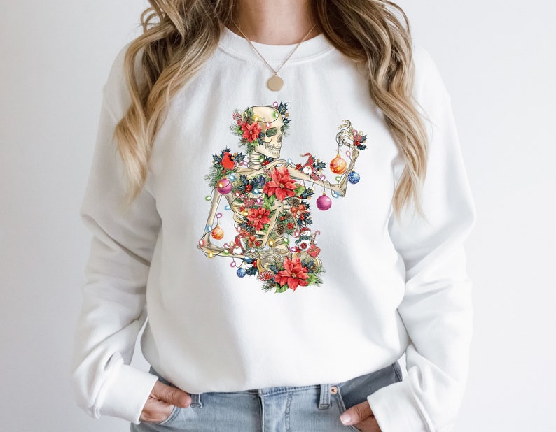 May include: White sweatshirt with a colorful illustration of a skeleton decorated as a Christmas tree. The skeleton is adorned with ornaments, lights, and poinsettias.