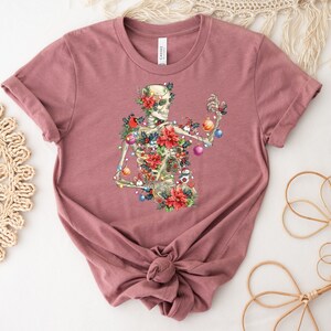May include: A mauve t-shirt with a skeleton wearing a Christmas wreath and holding ornaments. The skeleton is surrounded by poinsettias, holly, and other Christmas greenery.