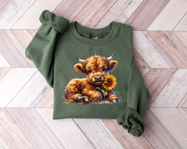 May include: A green sweatshirt with a brown and white Highland cow calf holding a yellow sunflower. The calf is sitting on a bed of green grass.