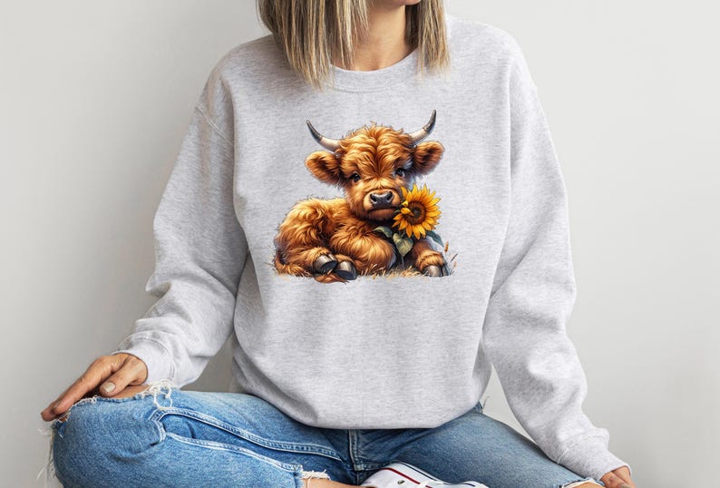 May include: A gray sweatshirt with a cute cartoon illustration of a brown Highland cow holding a yellow sunflower.