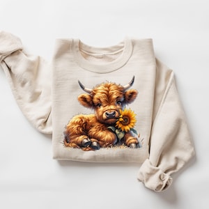 May include: A beige sweatshirt with a brown and white Highland cow calf holding a yellow sunflower. The calf is looking at the camera.