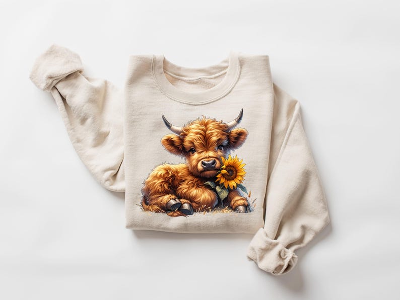 May include: A beige sweatshirt with a brown and white Highland cow calf holding a yellow sunflower. The calf is looking at the camera.