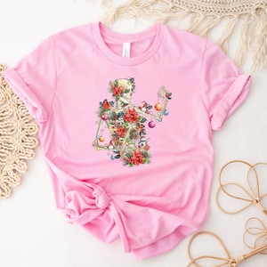 May include: Pink t-shirt with a skeleton decorated with Christmas ornaments, holly, and poinsettias.