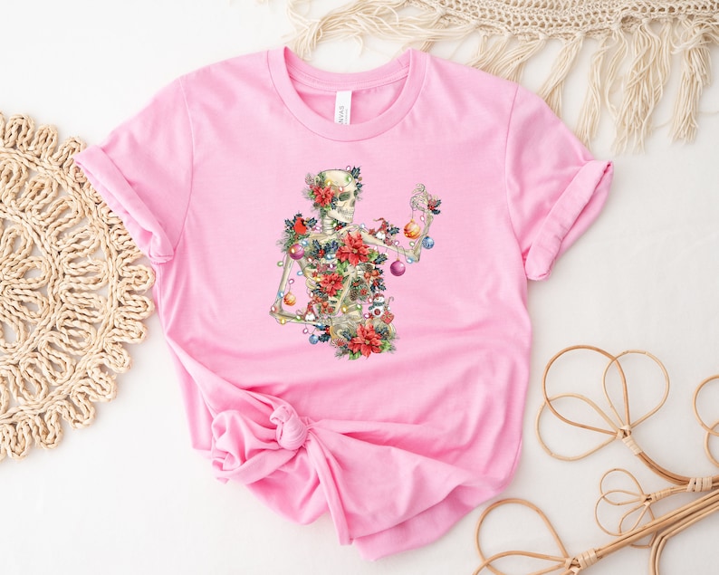 May include: Pink t-shirt with a skeleton decorated with Christmas ornaments, holly, and poinsettias.