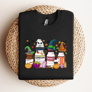 May include: Black t-shirt with a Halloween-themed graphic featuring three bottles of medication with witch hats and pumpkins. The bottles are labeled "Midazolam", "Diprivan", "Fentanyl", and "Alvan".