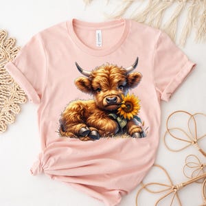May include: A pink t-shirt with a cartoon image of a brown Highland cow calf with a sunflower in its mouth.