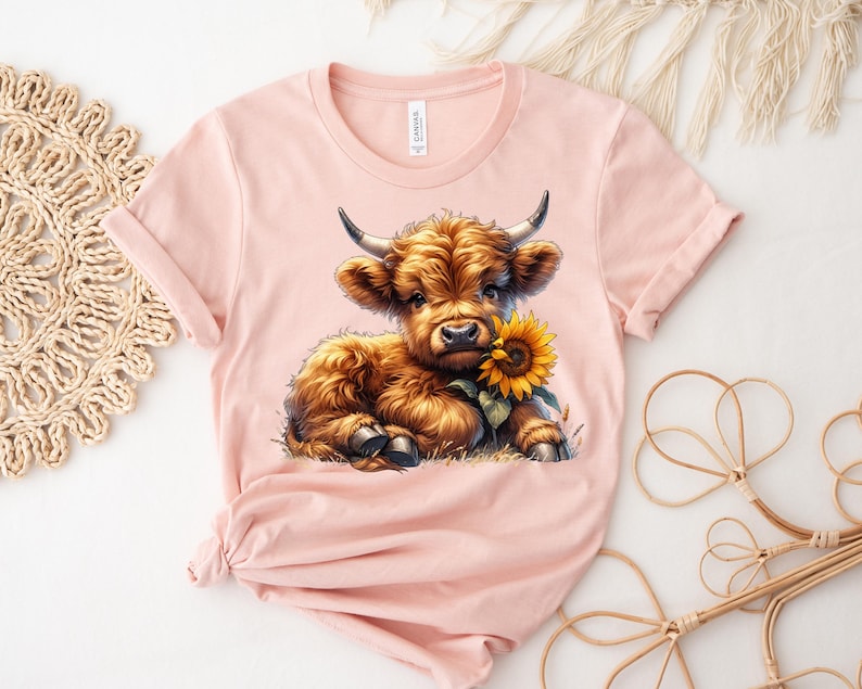 May include: A pink t-shirt with a cartoon image of a brown Highland cow calf with a sunflower in its mouth.