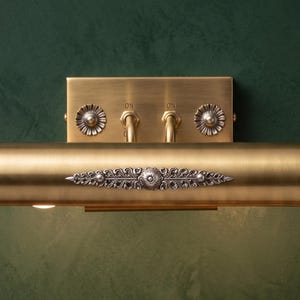 May include: A wall-mounted picture light with a brushed brass finish, featuring a cylindrical lamp body, a rectangular backplate, two toggle switches labeled ON, and decorative silver-toned floral accents on the center and sides.