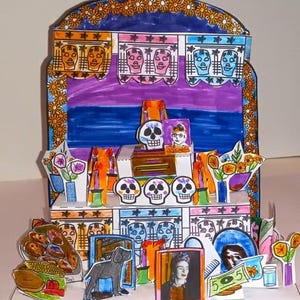 May include: A colorful, handmade Day of the Dead altar diorama. The artwork features painted skulls, flowers, and portraits, with a vibrant color palette of orange, purple, blue, and pink. The diorama is a festive and decorative piece.