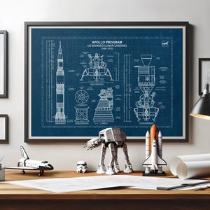 May include: A framed blueprint-style poster of the Apollo Program US Manned Lunar Landing (1960-1972). The poster is blue with white line drawings of the rocket and lunar module. Miniature models of a spaceship, AT-AT walker, and a space shuttle are on the desk.