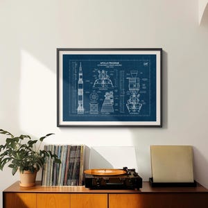 K&ouml;nnte beinhalten: Ein gerahmter Bauplan des Apollo-Programms, US Manned Lunar Landing (1960-1972), in Wei&szlig; auf dunkelblauem Hintergrund. Der Druck ist in einem schwarzen Rahmen. Der Druck h&auml;ngt &uuml;ber einem Holzschrank mit einem Plattenspieler, Schallplatten und einer Topfpflanze.