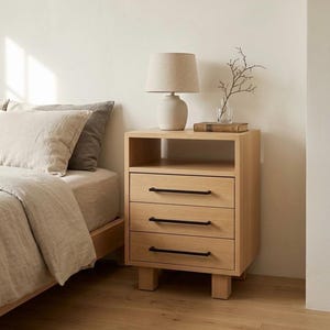 May include: A light wood nightstand featuring an open top shelf and three drawers with thin, horizontal black metal handles. The nightstand stands on four block feet and is topped with a beige table lamp and a small glass vase with a twig.