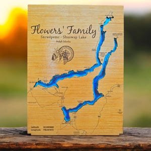 May include: A wooden map of Secwépemc - Shuswap Lake in British Columbia, with the words "Flowers' Family" at the top. The lake is outlined in blue and the map is on a wooden background.