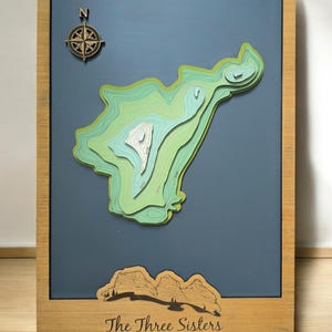 May include: A wooden framed map of The Three Sisters mountains in Canmore, Alberta, Canada. The map is a 3D topographic relief map with green and blue colours. The map is framed in a light brown wood frame.