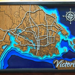 Puede incluir: Un mapa de madera enmarcado de Victoria, Columbia Británica, con un diseño en capas que muestra las calles y vías fluviales de la ciudad. El mapa presenta una rosa de los vientos y el nombre de la ciudad en cursiva. El agua está representada en tonos azules.