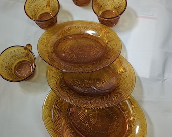 May include: Amber-colored glass dish set featuring floral and dot patterns. The set includes four handled cups and four oval plates. The dishes have a vintage aesthetic, perfect for serving meals or displaying as decor.
