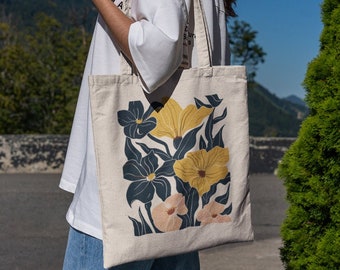 May include: A white canvas tote bag with a floral design featuring yellow, pink, and blue flowers on a dark blue background. The bag has two handles and is being held by a person.