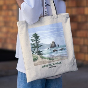 Può includere: Una borsa tote beige con un'illustrazione ad acquerello di Haystack Rock in Oregon. La borsa ha manici neri e il testo "Haystack Rock Oregon" stampato sul fondo.