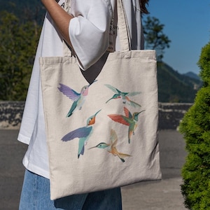 May include: A beige tote bag with a design of colorful hummingbirds. The bag has two straps and is made of canvas.