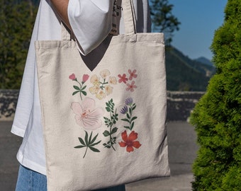 May include: A white canvas tote bag with a floral design featuring pink, red, yellow, and green flowers. The bag has two handles and is being held by a person.