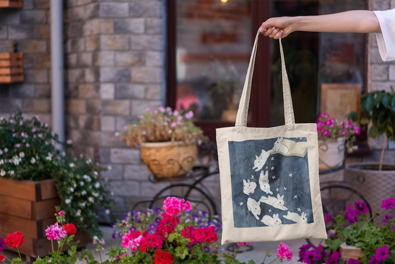 May include: A beige tote bag with a blue background and white rabbits jumping in the air. The rabbits are surrounded by white stars. The bag has two long straps.