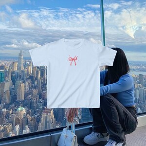 May include: A white t-shirt with a pink bow on the front. The shirt is being worn by a person standing in front of a window with a view of a city skyline.