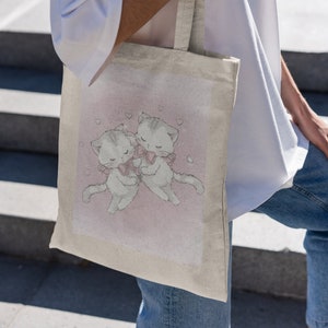 May include: A white canvas tote bag featuring a pink background with two cartoon cats wearing pink bows. The cats are hugging and there are small pink hearts around them.