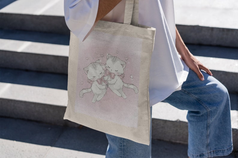 May include: A white canvas tote bag featuring a pink background with two cartoon cats wearing pink bows. The cats are hugging and there are small pink hearts around them.