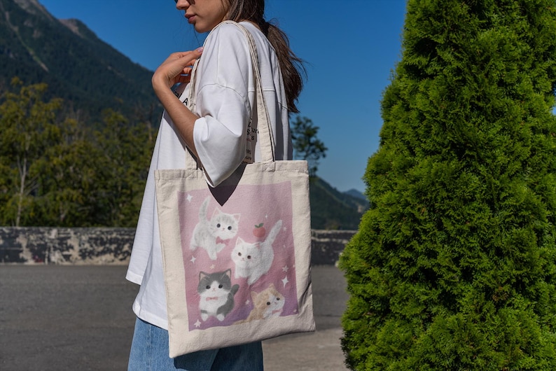 May include: A canvas tote bag featuring a pink background with illustrations of white and gray kittens. One kitten has a red bow tie and another has a strawberry on its head. The bag has a cream-colored handle.