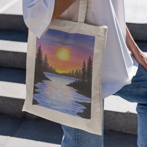 May include: A white canvas tote bag with a colorful sunset scene painted on it. The scene features a lake with trees on either side and a bright orange sun in the sky.