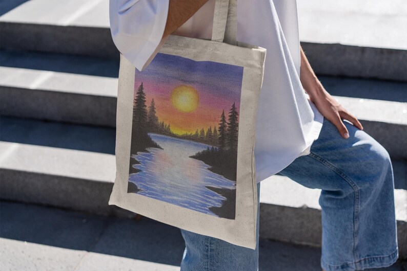 May include: A white canvas tote bag with a colorful sunset scene painted on it. The scene features a lake with trees on either side and a bright orange sun in the sky.