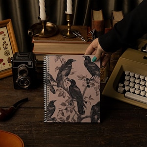Può includere: Un quaderno a spirale con una copertina rosa che presenta un'illustrazione in bianco e nero di corvi su un ramo. Il quaderno è su un tavolo di legno con una macchina fotografica vintage, una pipa e una stampa incorniciata di funghi.